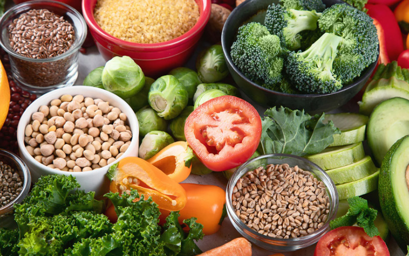 A imagem é composta por vários tipos de alimentos coloridos, como legumes, frutas, vegetais e grãos, por exemplo: brócolis, abacate, alface, tomate, arroz, grão de bico e limão.