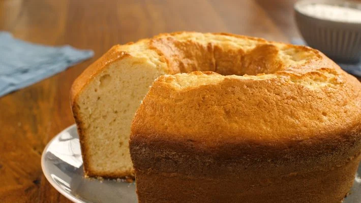Fotografia de um bolo redondo com um furo no meio, em um prato branco, sobre uma mesa de madeira. O bolo está bem cozido e dourado, cortado, faltando um pedaço.