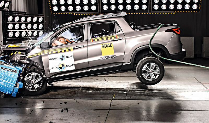 A imagem mostra um teste realizado com um veículo em alta velocidade. Um carro prateado choca-se contra uma parede, demonstrando a dispersão de energia na colisão.