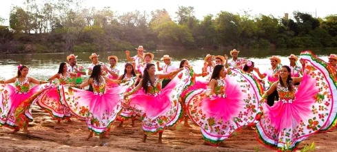 Imagem com cinco mulheres em destaque, dançando à margem de um rio. Estão usando vestido longo colorido rosa com a parte superior e a barra floridas. Ao fundo, há alguns homens de chapéu, dançando e aplaudindo a dança.