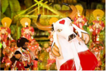 Imagem da Festa do Boi-Bumbá. No primeiro plano, consta o fantoche da cabeça de um boi branco, com chifres grandes cor de marfim, focinho preto e um coração vermelho desenhado na testa. Em segundo plano, de forma desfocada, aparece o restante da festa.