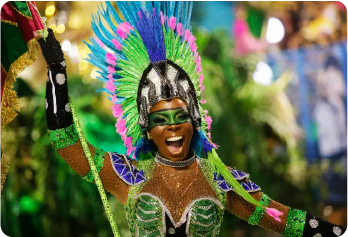 A imagem mostra uma mulher negra sorrindo com trajes de carnaval. Seu corpo está visível da região do tórax para cima. Ela está segurando com sua mão direita o mastro de uma bandeira. Seu braço esquerdo está aberto. Seus gestos indicam que está dançando e cantando. Sua vestimenta remete à cultura indígena. Ela está com uma faixa pintada de verde na região dos olhos e nariz e está vestindo um cocar indígena repleto de penas coloridas nas tonalidades verde, roxo, rosa e azul.