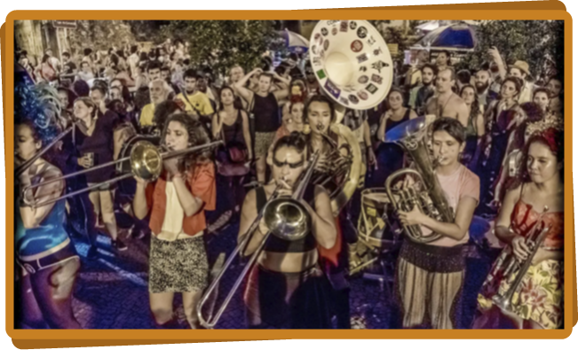 Foto de uma banda de fanfarra tocando na rua, com árvores e prédios ao fundo e diversas pessoas assistindo. Em primeiro plano, a banda é composta de seis mulheres tocando instrumentos de sopro, como trompete, tuba, clarinete e trombone, todas vestidas com roupas coloridas, tipo carnaval.