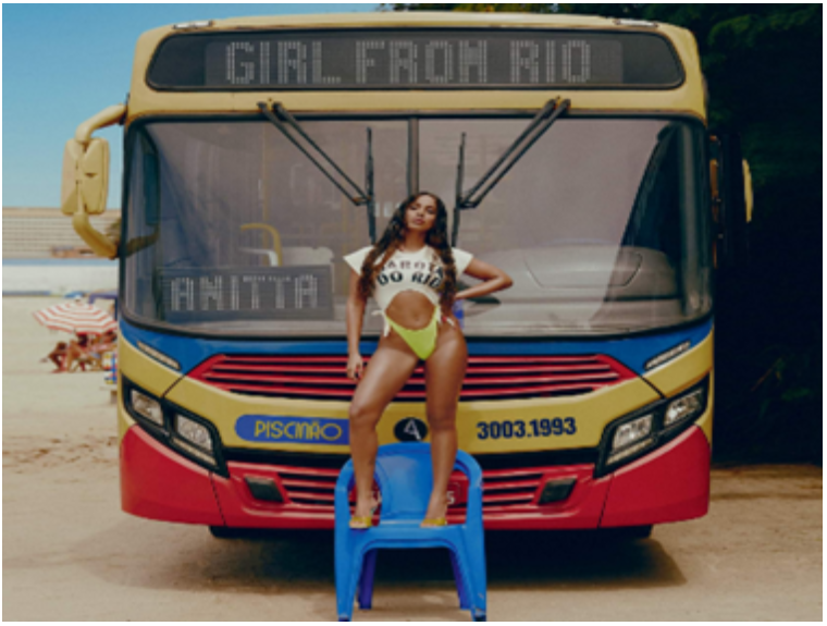 Fotografia da cantora Anitta em pé, em cima de uma cadeira, na frente de um ônibus, com o letreiro “Girl from Rio”. 