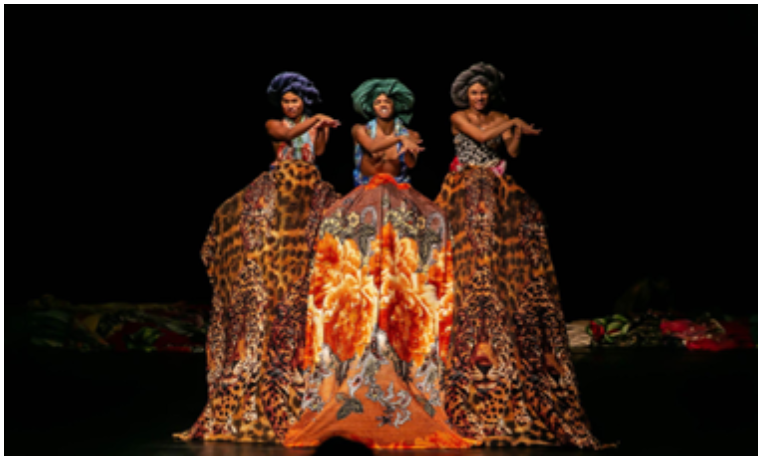 Três mulheres vestidas com saias longas e coloridas, em um palco. 