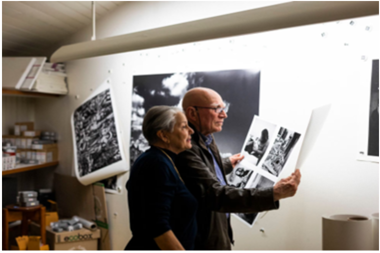 Fotografia do casal Sebastião Salgado e Lélia Wanick Salgado, dentro de uma sala, com diversos materiais de arte e fotografias. O casal está segurando e olhando fotografias em preto e branco. 