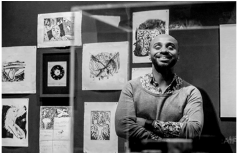 Fotografia de Igor Ramos, em preto e branco, em uma exposição de artes visuais. Igor está posicionado à frente de algumas obras, sorrindo, de braços cruzados. 