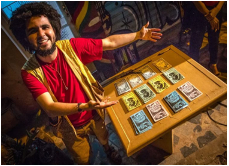 Diversos folhetos em papel colorido de literatura, posicionados lado a lado, em uma bancada de madeira. Ao lado da bancada, há um homem sorrindo e mostrando os folhetos. 